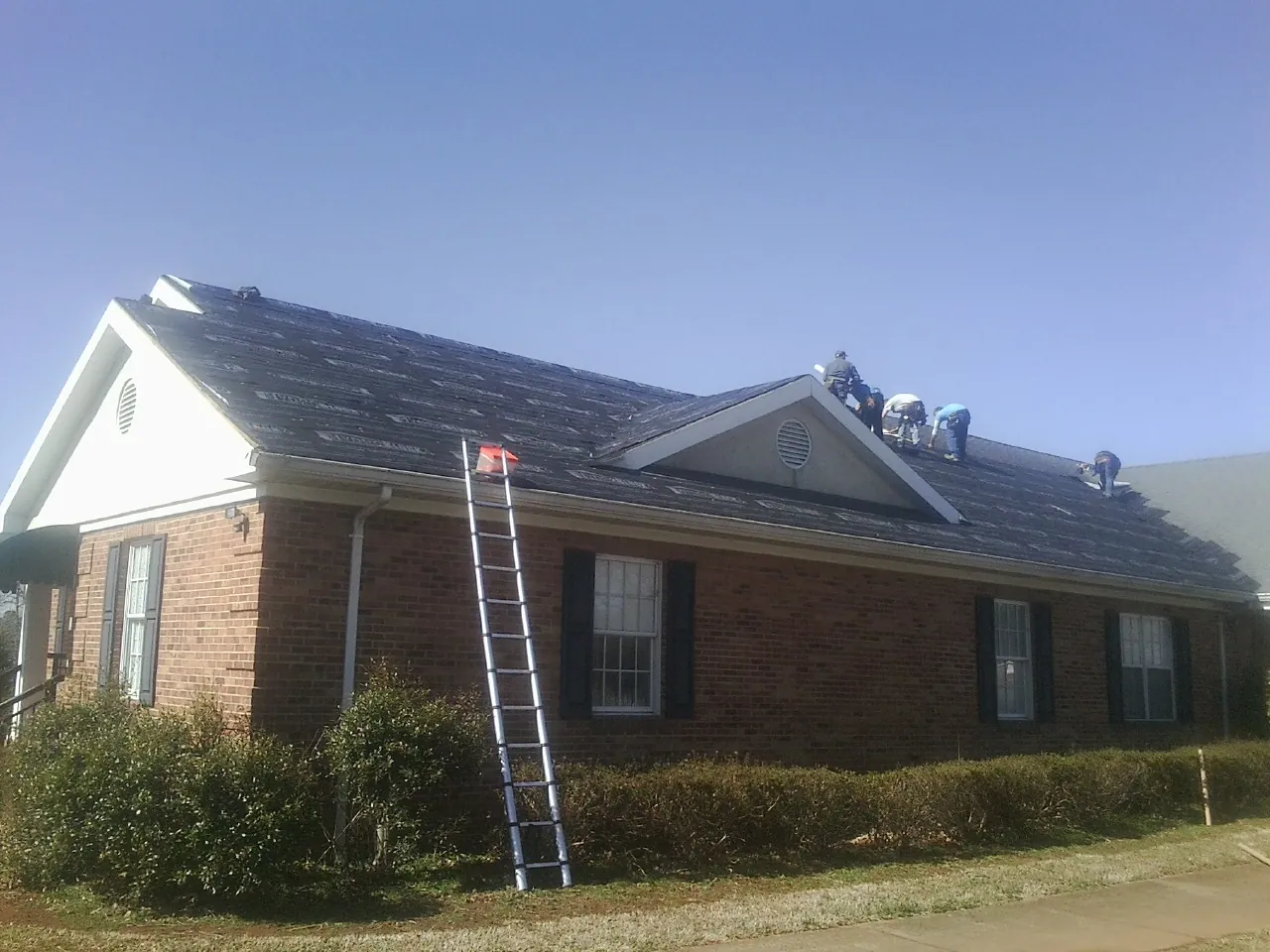 Crew tearing shingles off a brick ranch roof with a ladder set — Emergency Roof Repair work by our crew in Gnadenhutten