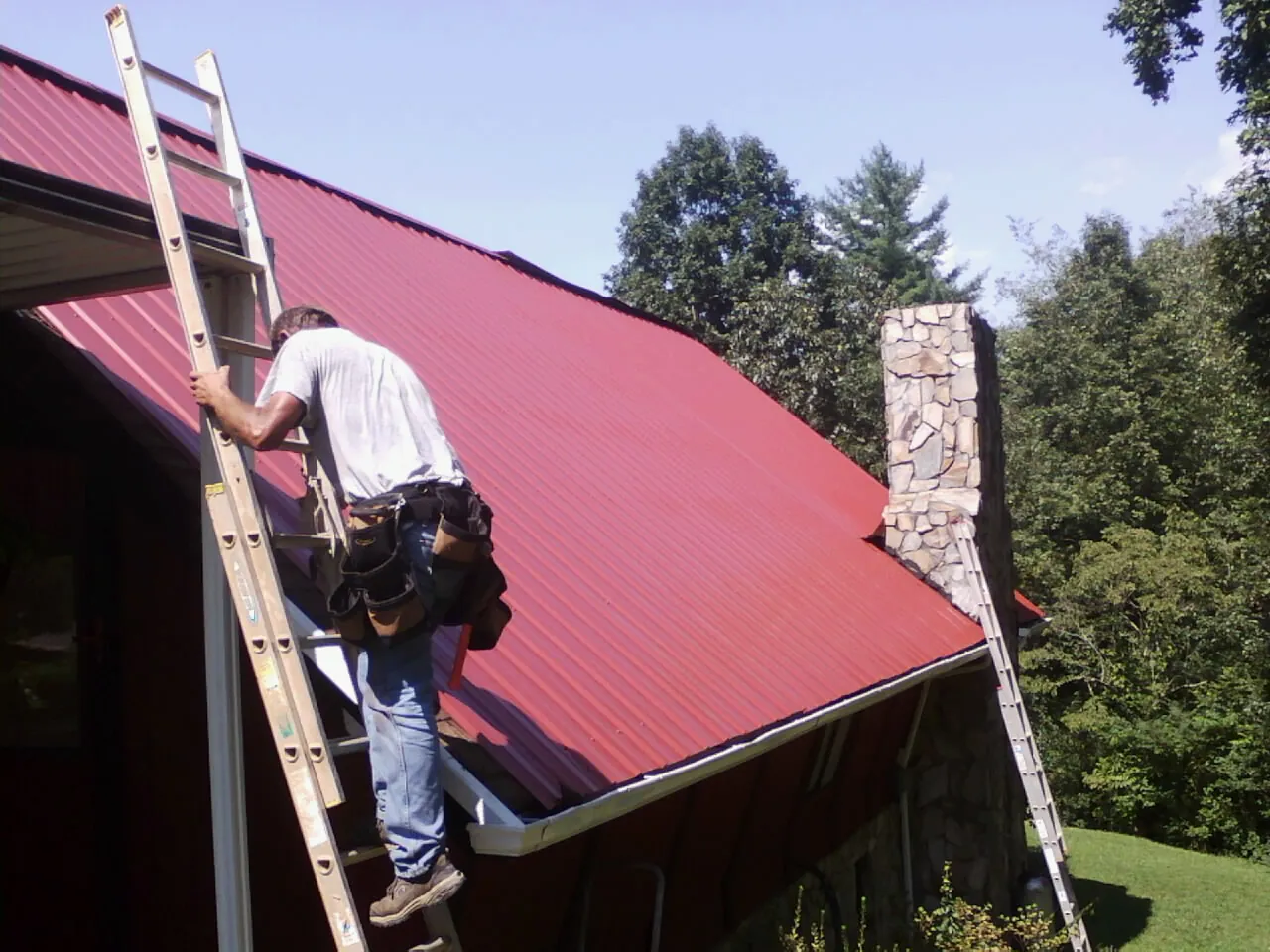 Roofer on a ladder beside a red metal roof and stone chimney — Soffit & Fascia Repair by Roofing Brothers in Gnadenhutten